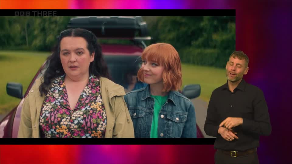 Two women stand next to a car, their faces turned towards each other. A man on the right signs along to the programme on BBC Three.
