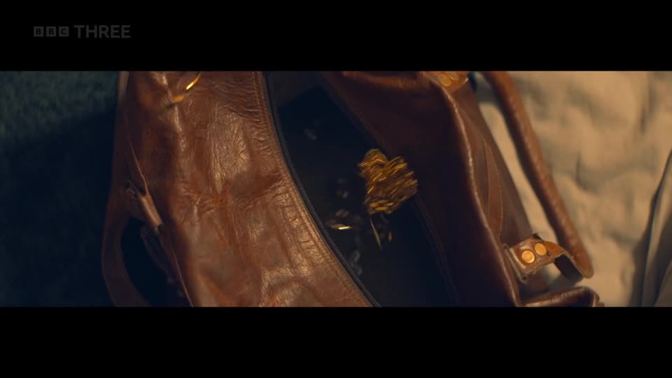 A brown leather bag lies open, its contents glinting under the light. A small, golden object, like a crumpled foil flower, sits atop a pile of what appears to be coins.