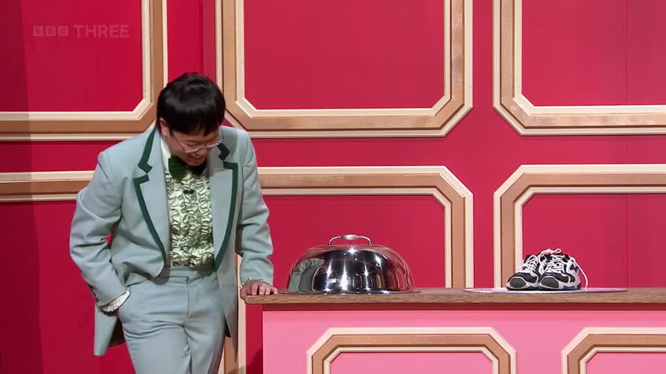 A man in a light blue suit with green trim leans over a counter. On the counter, a silver cloche sits next to a pair of black and white trainers. The BBC Three logo is visible in the top left corner.