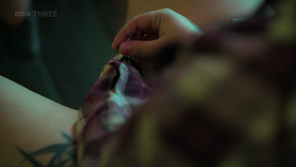 A hand is carefully threading a needle through a purple and white checkered fabric. The BBC Three logo is visible in the top left corner.