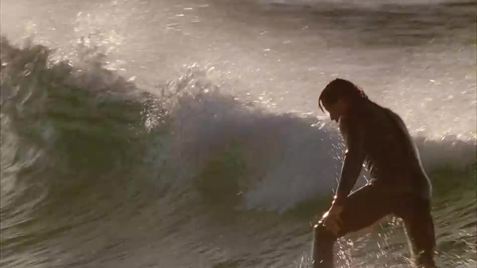 A surfer in a dark wetsuit carves through a breaking wave. The water sprays up around their board as the BBC Three cameras capture the moment.