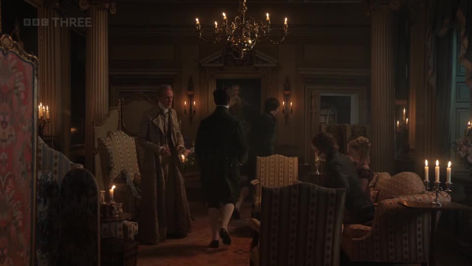 A man in a long coat walks across a dimly lit room, holding a candelabra. Several people are seated on ornate furniture, their faces obscured by shadow.