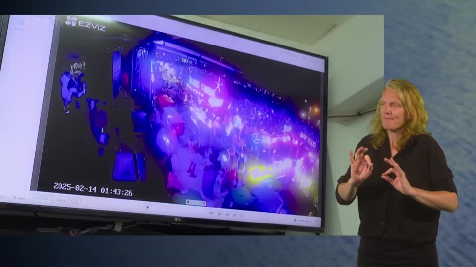 A crowd surges under bright, flashing lights, captured by a security camera. A woman signs beside the screen, her hands moving with the rhythm of the broadcast.