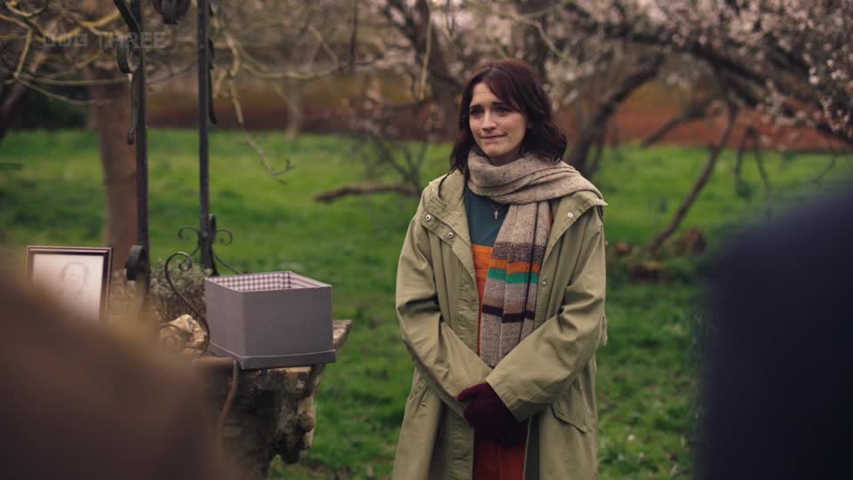 A young woman stands in a garden, clutching her hands together. A framed photograph and an empty box sit on a stone pedestal beside her.
