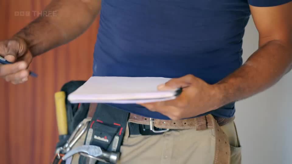 A man in a blue t-shirt and tool belt holds a notepad and pen. He's about to write something down.