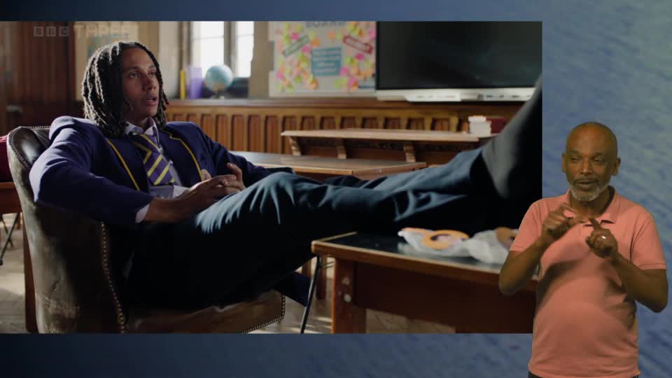A young man in a school blazer relaxes with his feet up on a desk. To the right, a man signs with his hands.