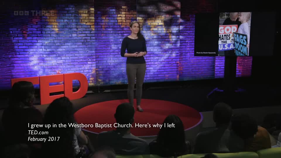 A woman stands on a red circular stage, speaking into a microphone. Behind her, the large red TED logo is visible, and a screen displays an image of a protest with signs. This is a BBC Three broadcast from the UK.