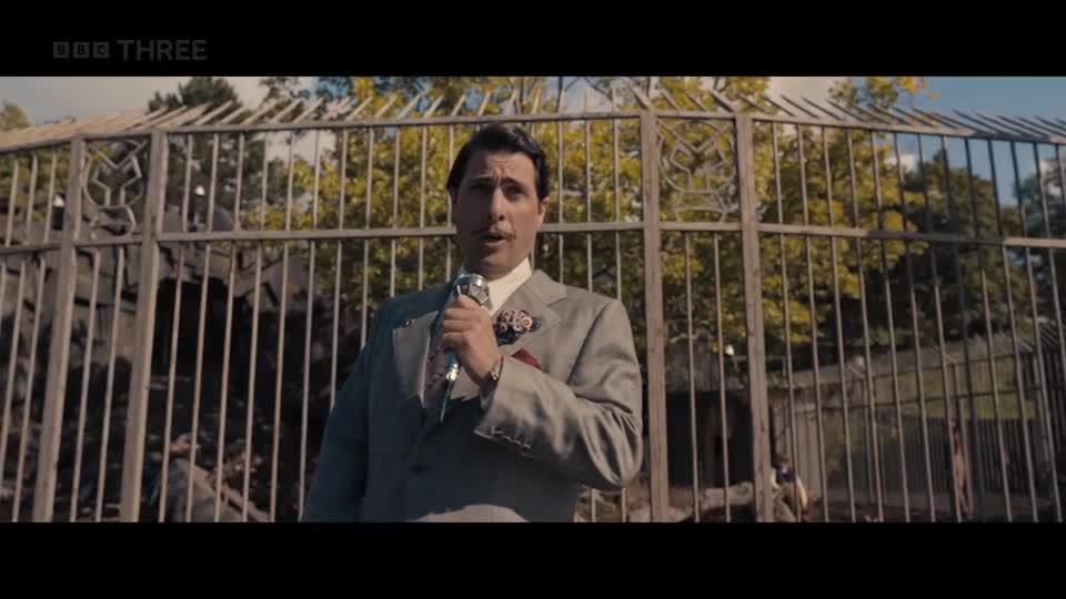 A man in a sharp grey suit and tie speaks into a microphone, his expression animated. Behind him, a large, ornate metal gate stands open, revealing trees and a glimpse of a building. A man in a sharp grey suit and tie speaks into a microphone, his expression animated. Behind him, a large, ornate metal gate stands open, revealing trees and a glimpse of a building.