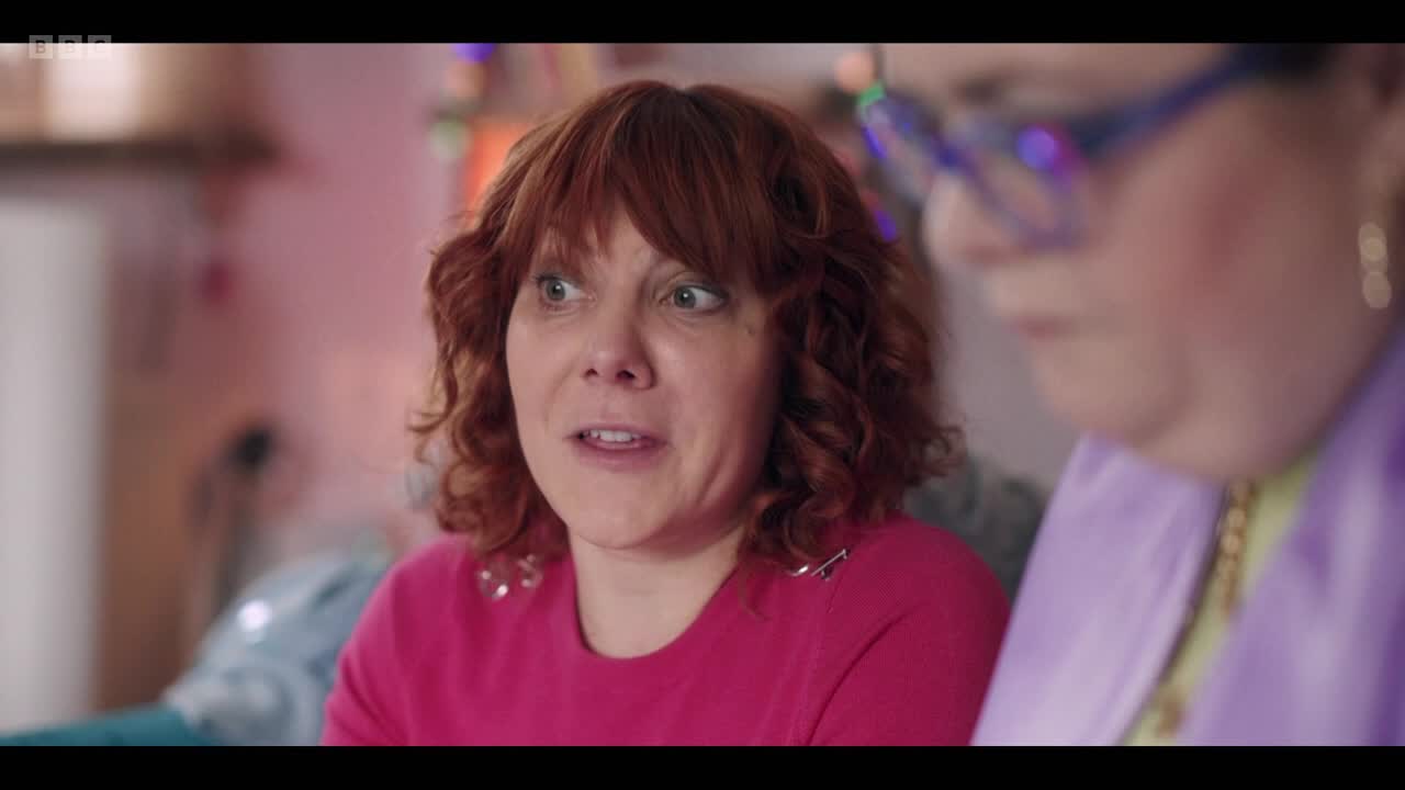 The woman in the pink sweater is talking, her eyes wide with surprise. Beside her, another woman in a lilac jacket listens intently, her face partially obscured.
