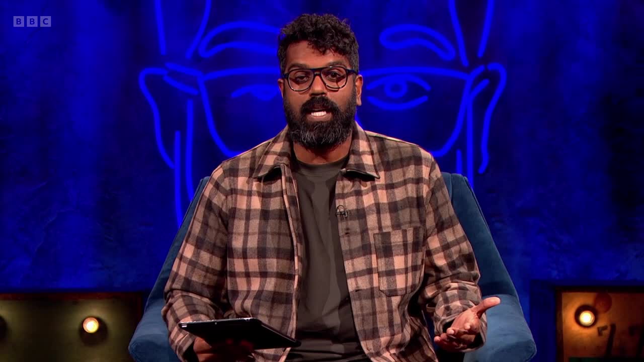 He's seated in a blue armchair, speaking, gesturing with his hands, and holding a tablet. Behind him, a neon-lit outline of a face glows against a dark blue backdrop, likely on BBC Two.
