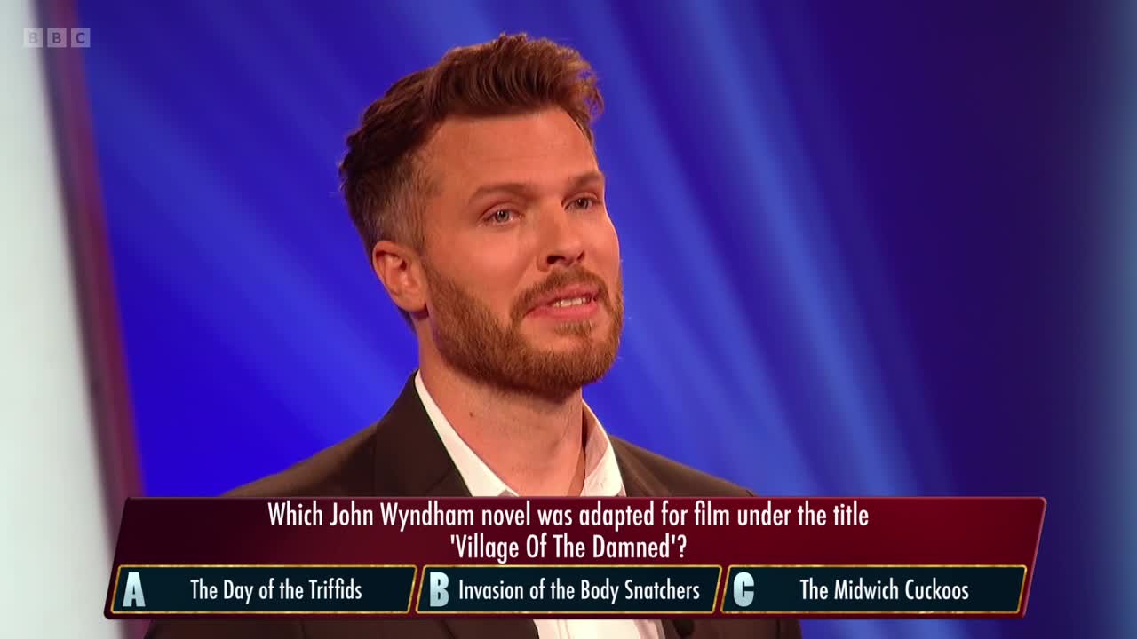 A man in a suit jacket is looking towards the camera, a slight smile on his face. Below him, a question about a John Wyndham novel appears on a screen, with three possible answers.
