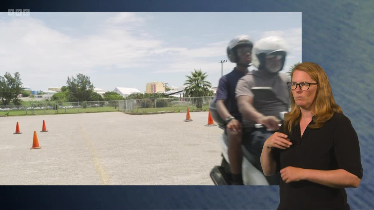 A scooter with two riders, both wearing helmets, is moving across a paved area. The presenter on BBC Two is signing, while the scooter appears to be navigating around orange cones.
