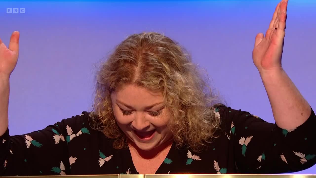 Her arms are raised, palms open, and she's looking down with a big smile. The woman's curly blonde hair frames her face as she appears to celebrate something.

