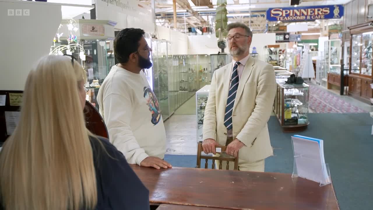 The man in the light suit stands near a small wooden chair, talking with the man in the sweatshirt. A woman with long blonde hair is behind the counter, listening.
