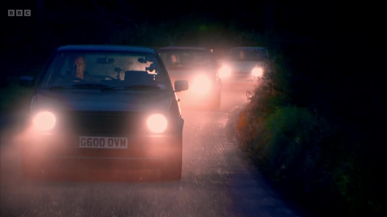 Headlights pierce the darkness as three cars drive toward me, their beams reflecting off the wet road. The first car's number plate is visible, and the driver is looking straight ahead.
