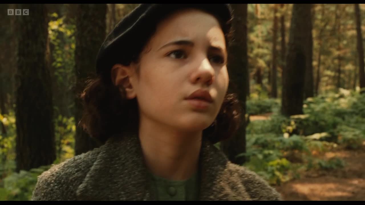 A young woman in a beret and textured coat looks off to the side, her expression thoughtful. Behind her, the trees of a forest fill the background.
