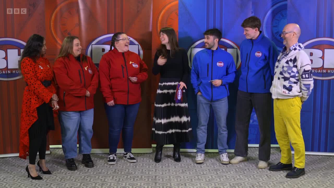 A group of people, some in red and blue jackets, are standing together on a set. The host, holding a card, chats with the contestants on the BBC Two show.
