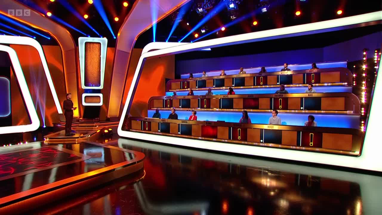 On a brightly lit stage, a man in a dark suit stands speaking, while contestants sit in tiered seating. The set design features large exclamation marks and a sleek, modern aesthetic.
