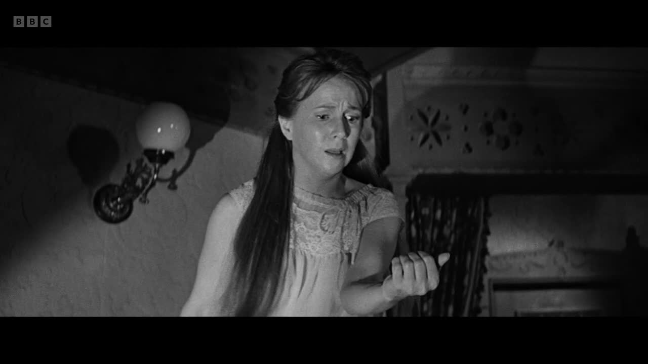 A woman in a nightgown looks down at her hand, her face etched with concern. The dim light from a wall sconce illuminates her expression as she seems to consider something.
