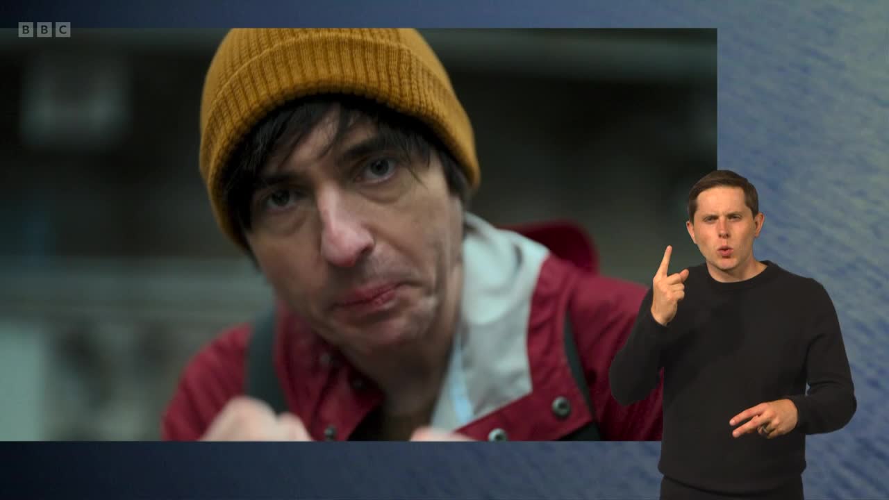 A man wearing a mustard-colored beanie stares directly at the camera, his lip slightly bloodied. Beside him, another man is signing, his hands moving in front of a blue backdrop.
