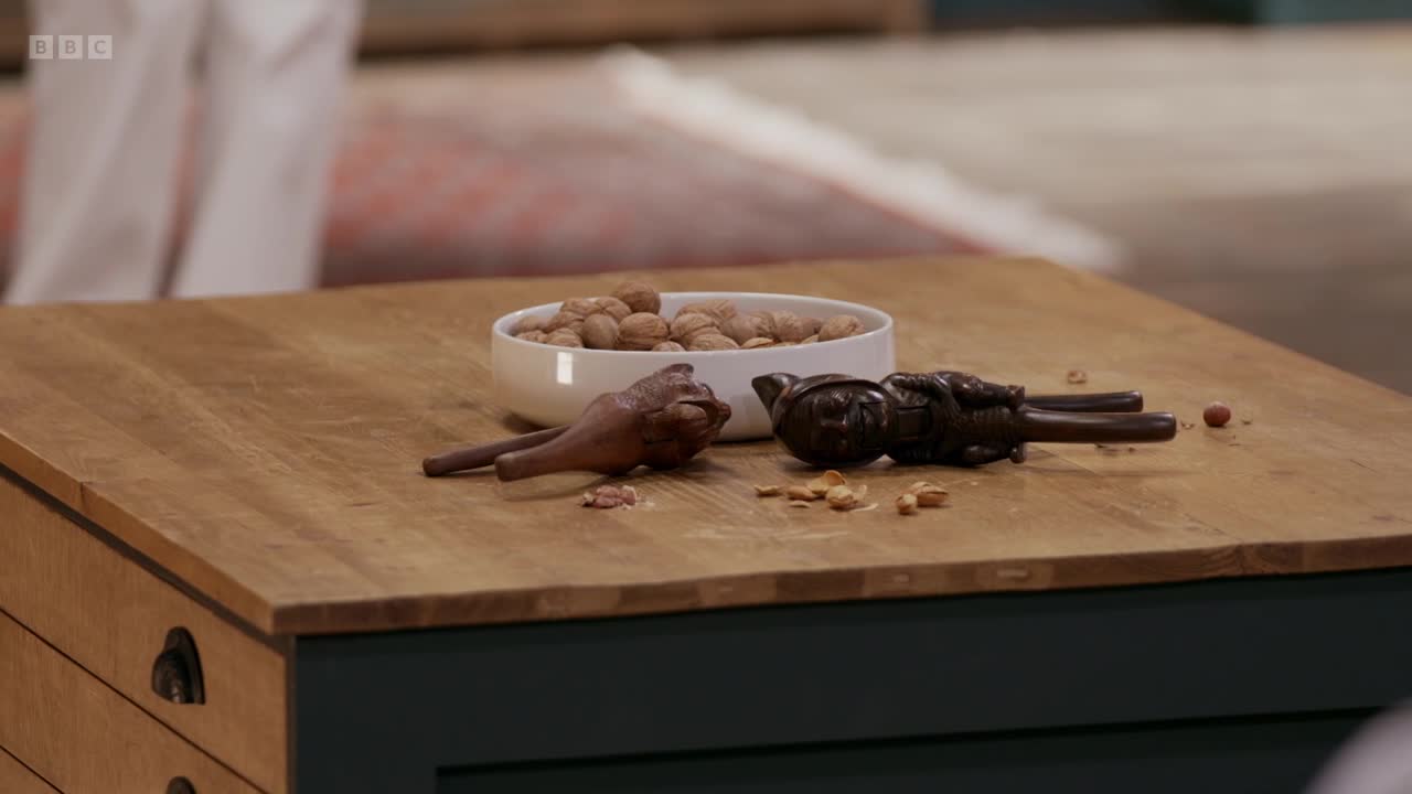 Two nutcrackers, one shaped like a frog and the other a man, lie open on a wooden surface scattered with shells and a few whole nuts. A white bowl overflowing with walnuts sits nearby.
