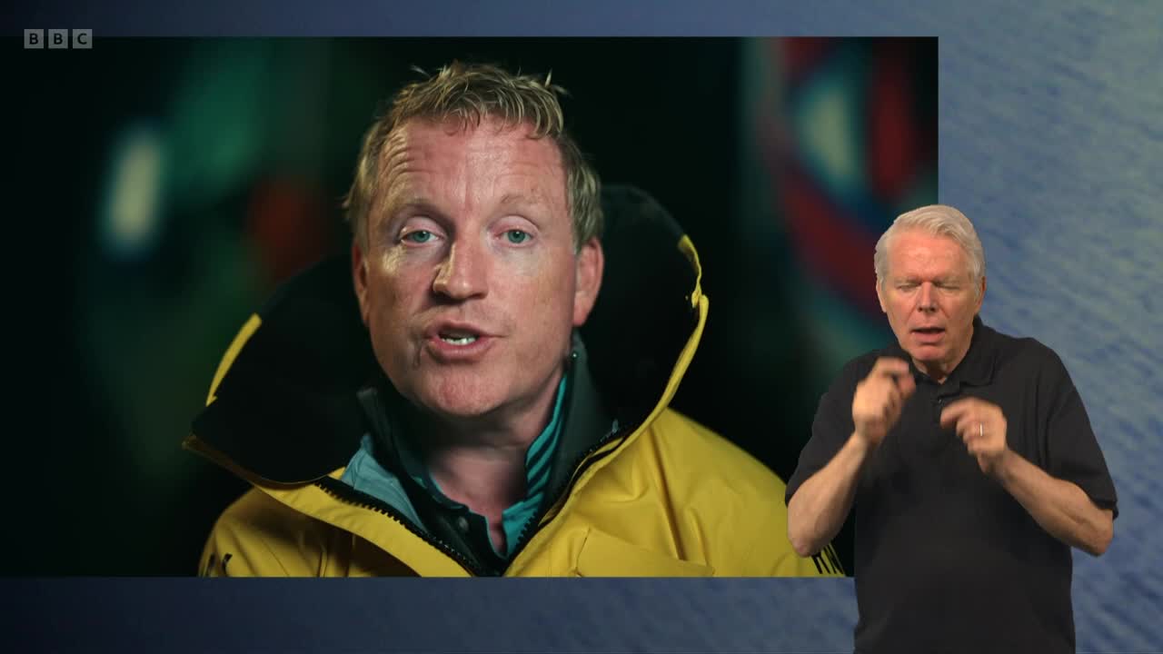 A man in a yellow jacket is speaking, his face filling the screen. Beside him, another man interprets his words with hand gestures.
