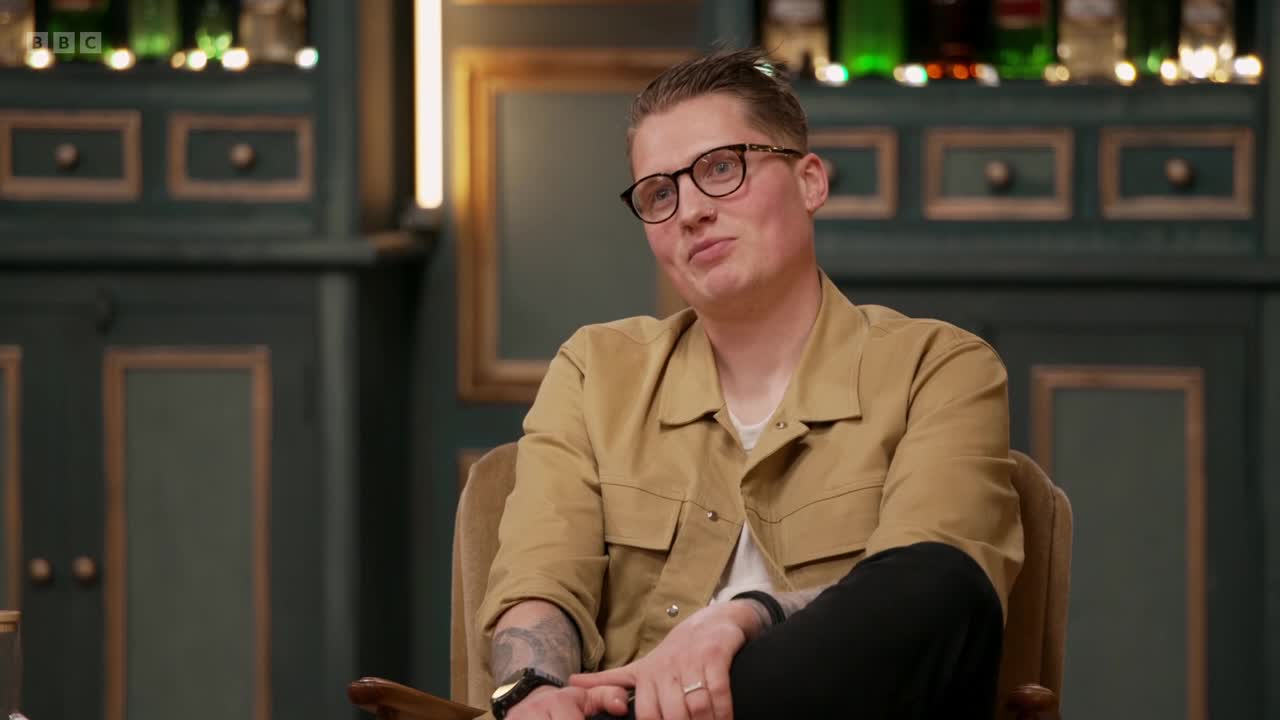 A man in a tan jacket and glasses sits in a chair and looks towards the camera with a slight smile. Behind him, the set of a BBC Two show in the United Kingdom is visible, with dark green paneling and framed artwork.
