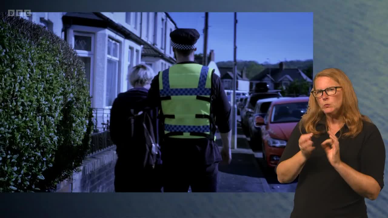 A police officer in a high-visibility vest walks down a residential street alongside someone with light-colored hair. The person on the right is signing.
