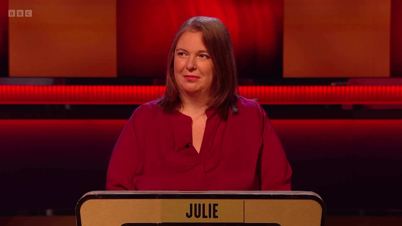 Julie, wearing a burgundy shirt, is at a podium with her name on it. She's looking forward, a slight smile playing on her lips under the studio lights.
