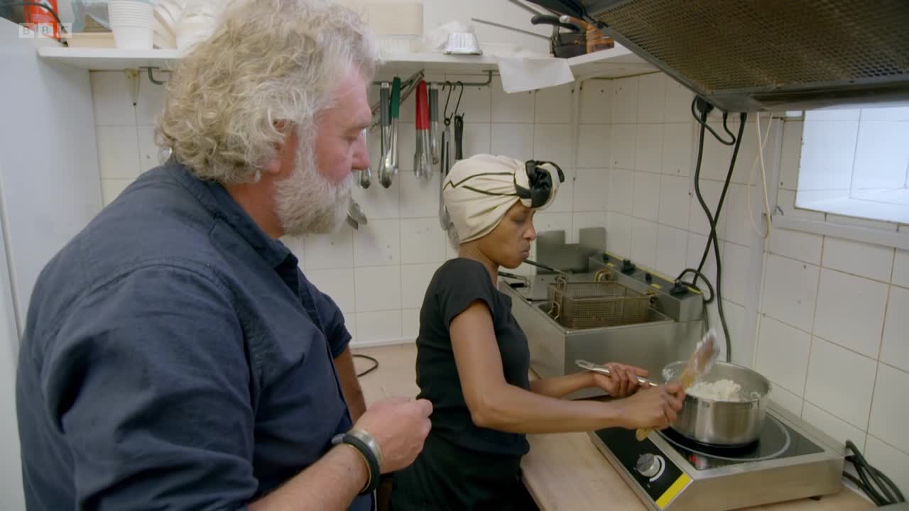A woman in a headscarf stirs something in a pot on a small electric hob. A man with a long white beard stands beside her, watching.
