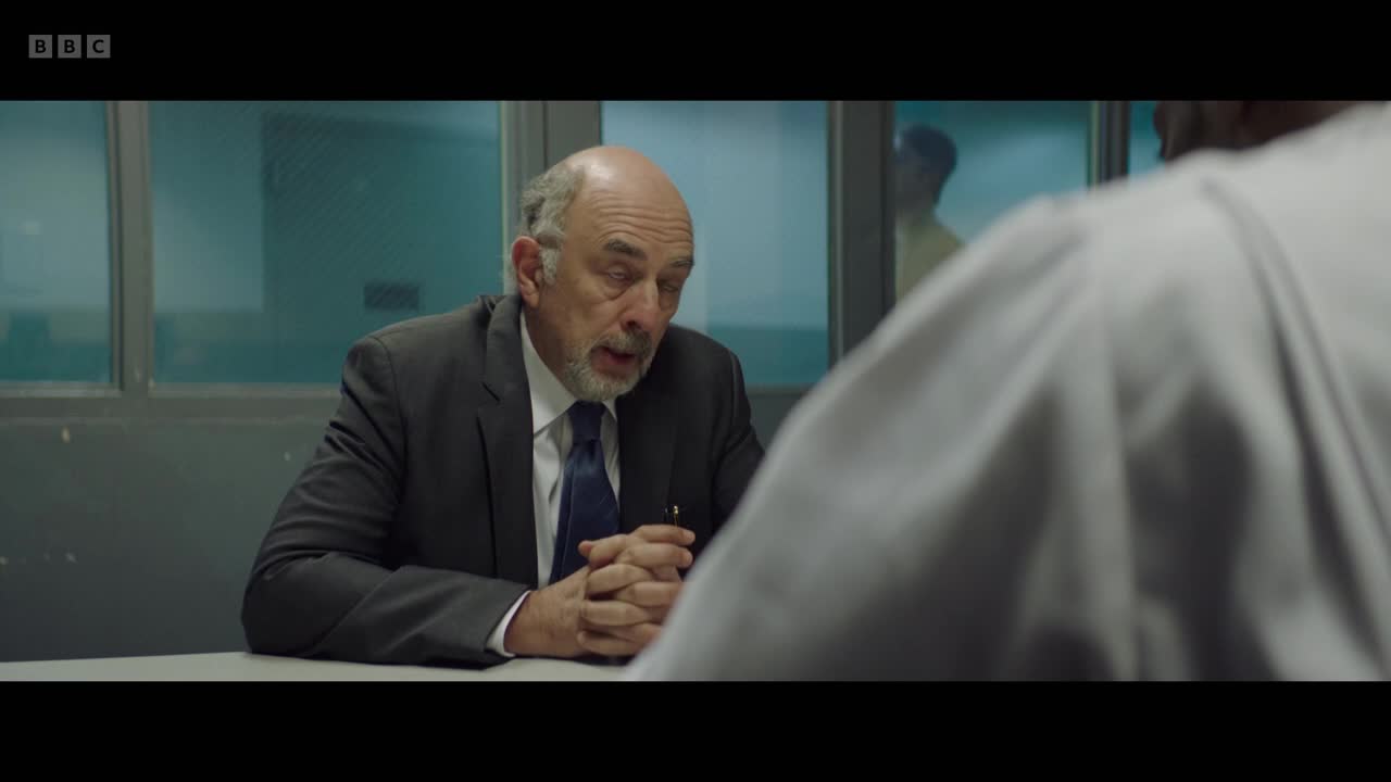 A man in a suit, hands clasped on the table, leans forward, speaking intently. Through the glass, another figure is visible, standing in what appears to be a holding cell.
