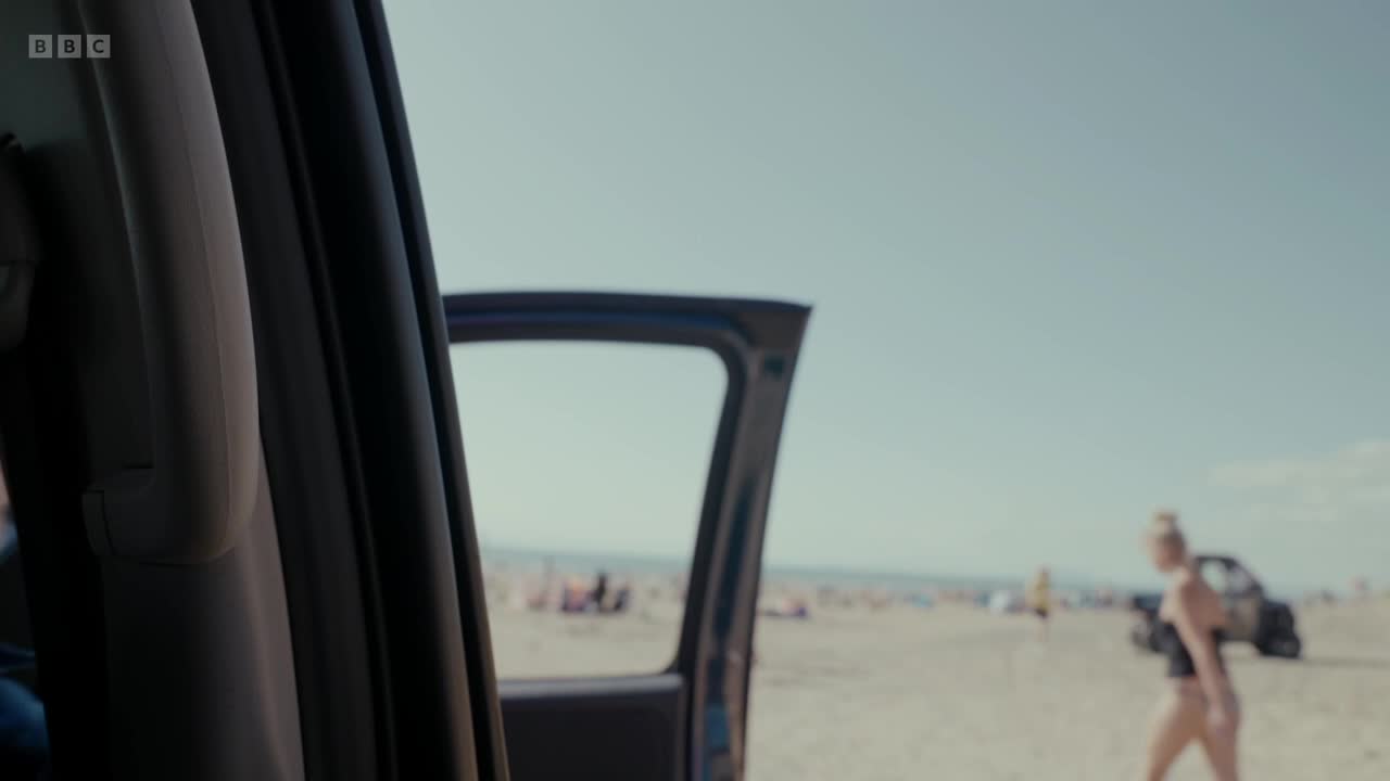 A woman in a black swimsuit walks across the sandy beach, heading away from the open car door. Beyond her, the beach is filled with people enjoying the sun.
