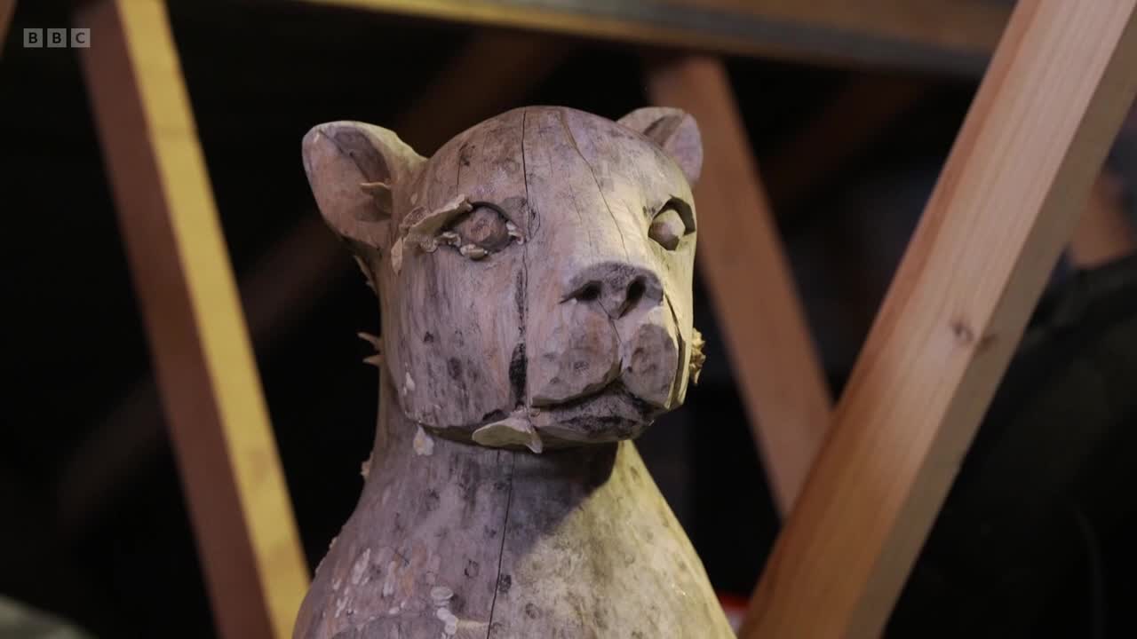 A carved wooden animal head, possibly a big cat, is the focus. The sculpture is set against a backdrop of wooden beams, likely in a workshop.
