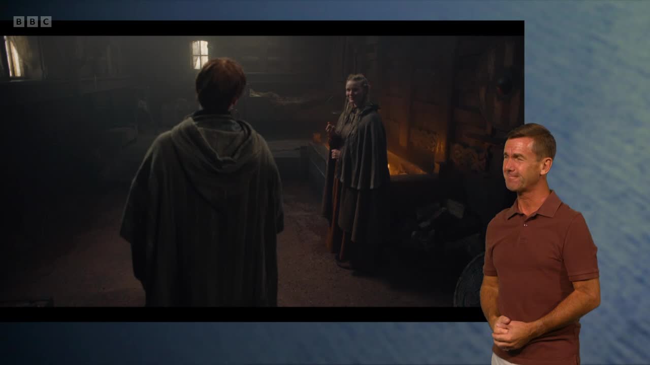 A figure in a hooded cloak faces two women in a dimly lit room. The woman on the right holds a staff, while the other waits, their backs to the camera.
