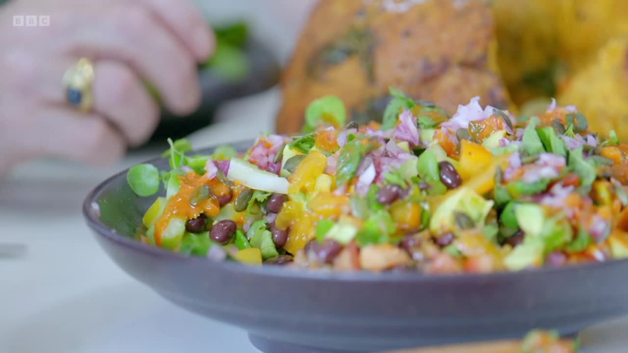 A vibrant salad, speckled with pumpkin seeds and black beans, sits in a dark bowl. In the background, a hand gently places more greens onto the dish, as seen on BBC Two.
