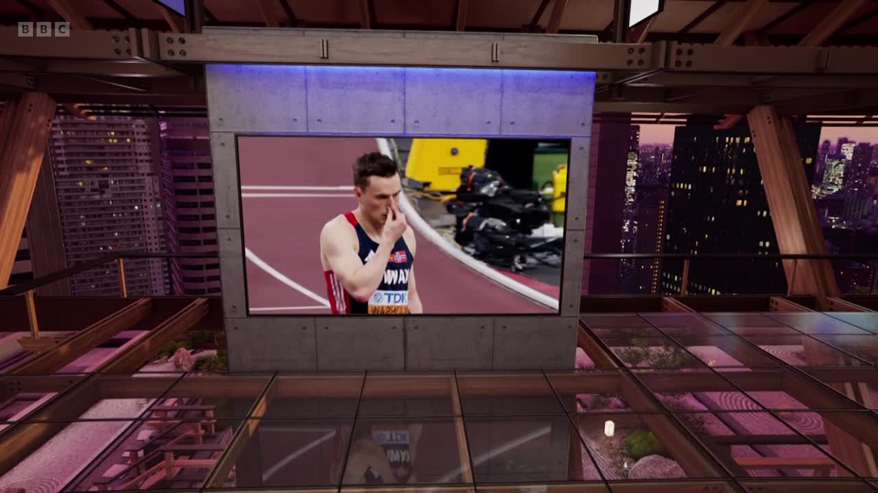 A large screen displays a track athlete in a Great Britain uniform, touching his nose. The BBC Two studio set features a glass floor and a cityscape backdrop.
