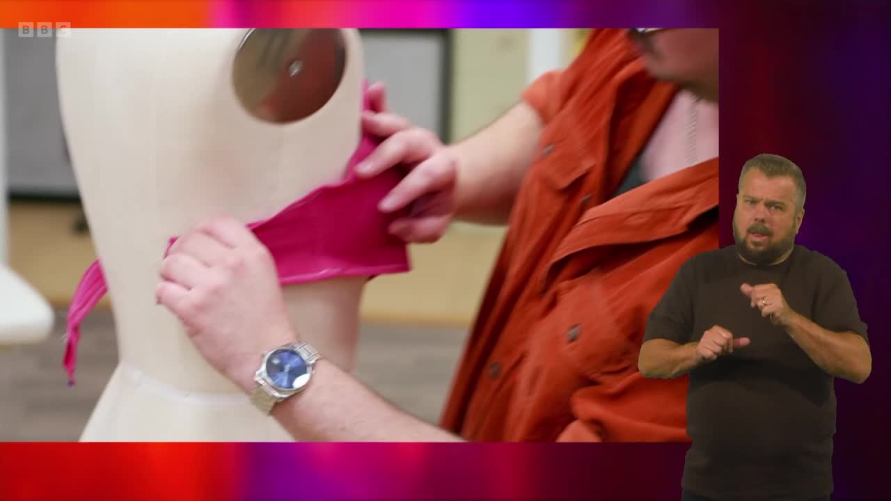 A person in an orange shirt is carefully draping a strip of pink fabric around a mannequin. Across the screen, a man in a black shirt is signing.
