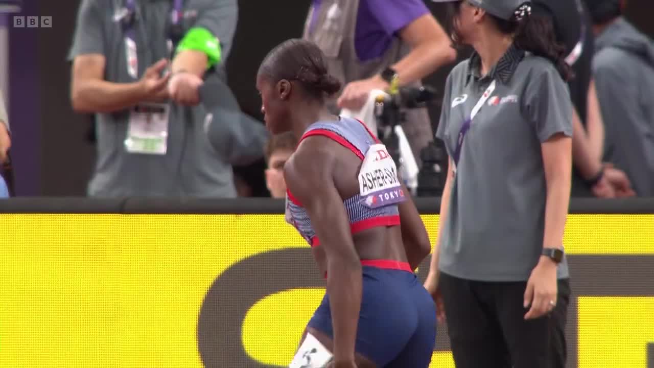 Dina Asher-Smith, wearing the British kit, is bent over, seemingly catching her breath after a race. A woman in a grey shirt and cap stands nearby, looking on.
