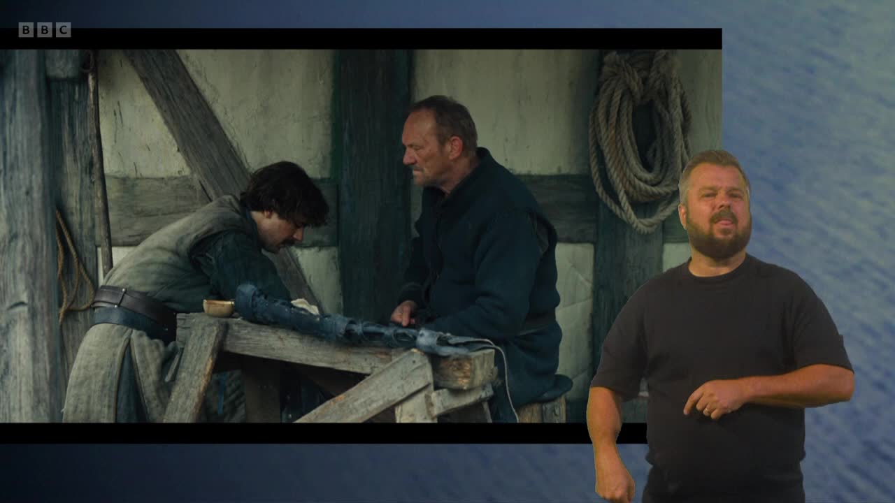 A young man leans over a workbench, working on a long, dark object. Beside him, an older man watches intently, while a sign language interpreter gestures in the foreground.
