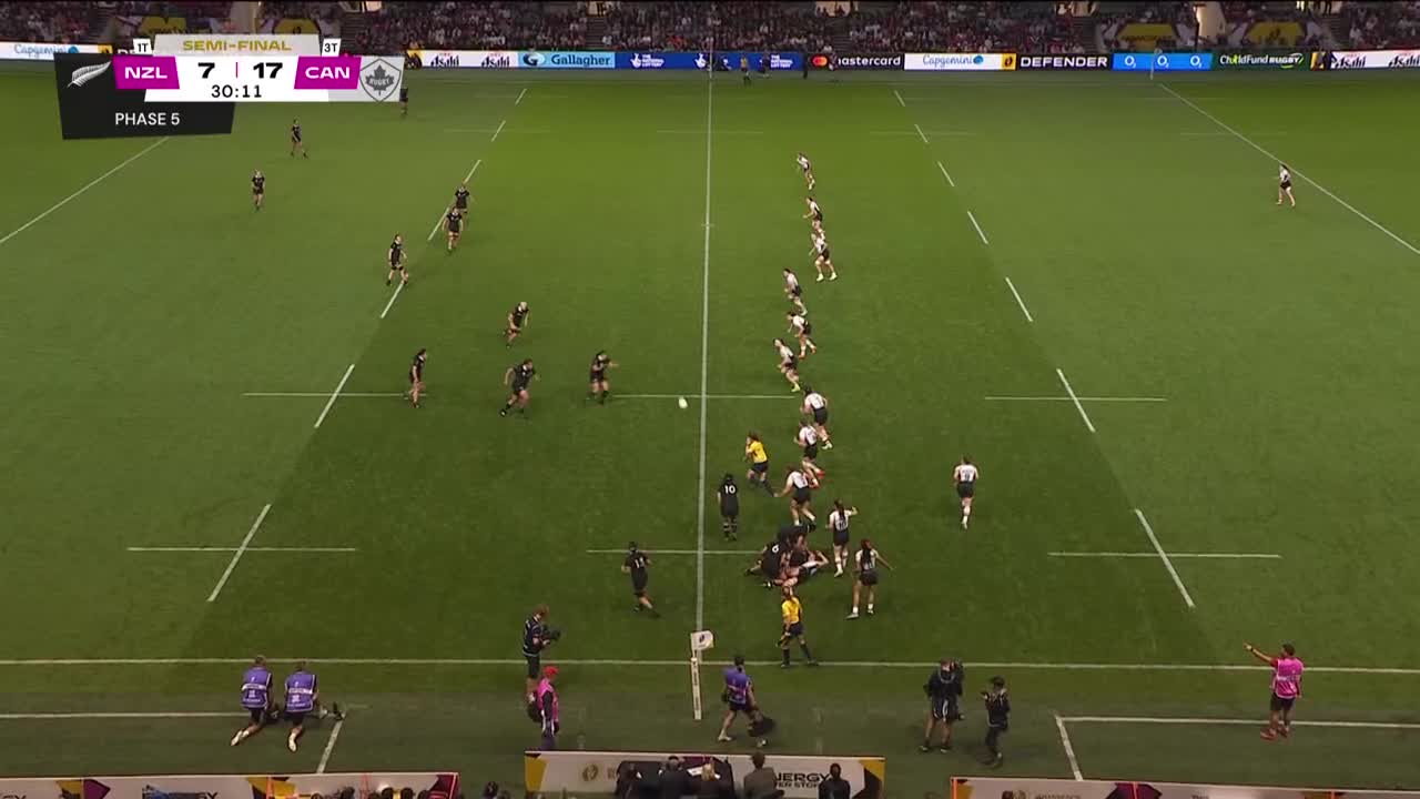 The ball hangs in the air as a scrum of players from both teams battle for possession. The referee, in yellow, watches closely as the play unfolds.
