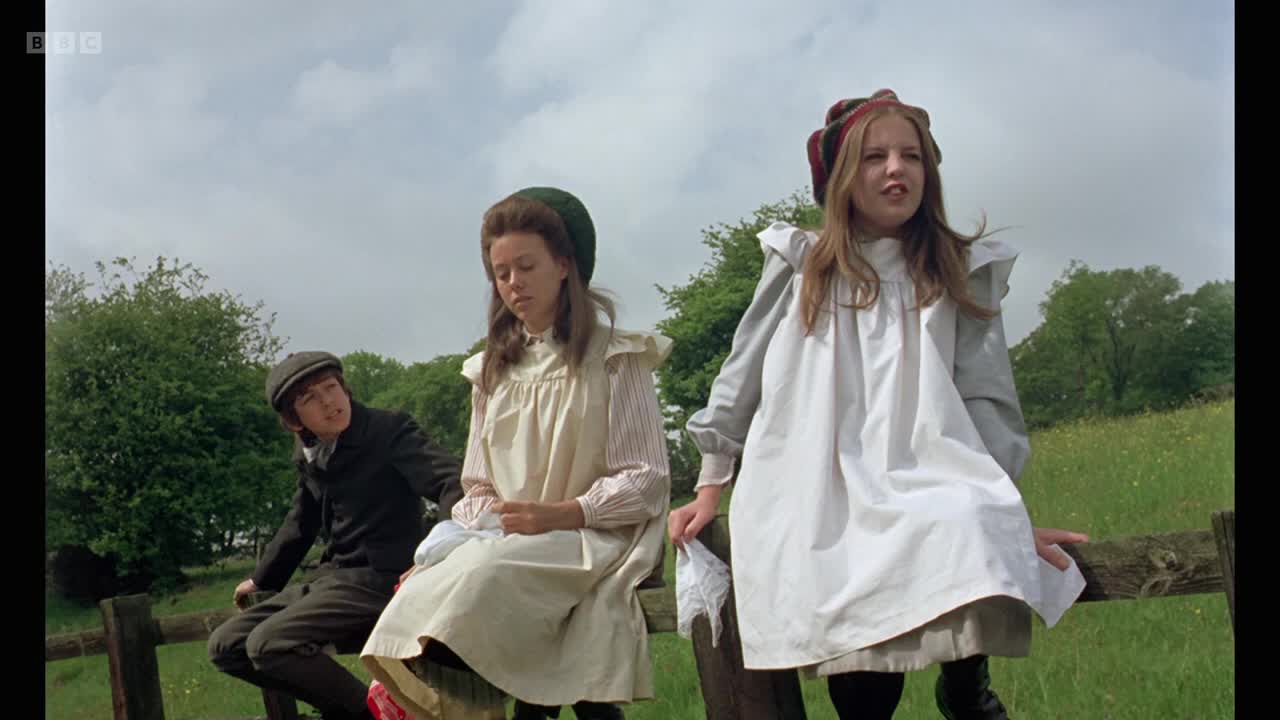 Three children are perched on a wooden fence, the girls in white dresses and bonnets, the boy in a cap and dark suit. The girl on the right appears to be speaking, while the other two are listening.
