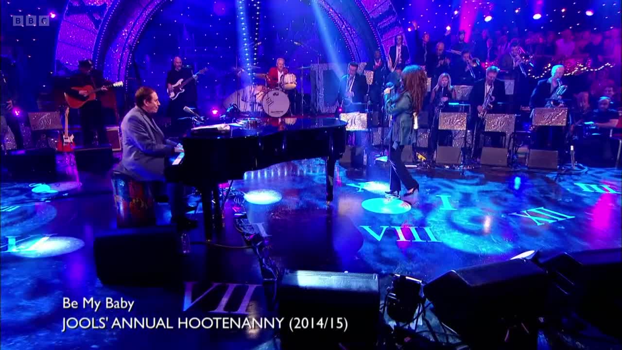 A pianist in a suit plays a grand piano, while a woman in a green jacket sings into a microphone. Behind them, a full band and a string section fill the stage, all under bright stage lights.
A pianist in a suit plays a grand piano, while a woman in a green jacket sings into a microphone. Behind them, a full band and a string section fill the stage, all under bright stage lights.