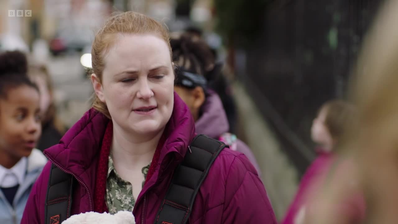 A woman in a burgundy jacket is talking, her face showing a mix of emotions. Children are visible in the background, and the scene feels like it's taking place outdoors.
A woman in a burgundy jacket is talking, her face showing a mix of emotions. Children are visible in the background, and the scene feels like it's taking place outdoors.