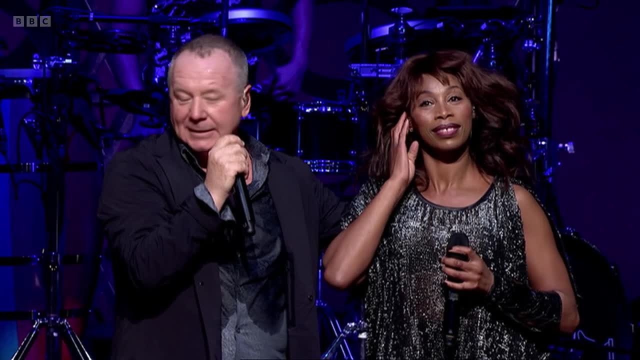 The man on the left holds a microphone, eyes closed, singing. Beside him, a woman with long hair gently touches her ear, also holding a microphone.
The man on the left holds a microphone, eyes closed, singing. Beside him, a woman with long hair gently touches her ear, also holding a microphone.