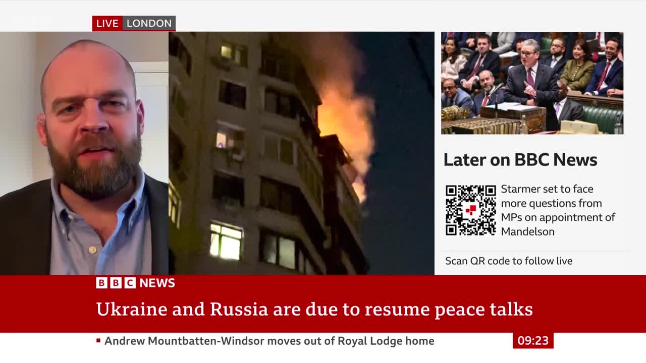 Flames erupt from an apartment building, casting an orange glow against the night sky. A man with a beard speaks directly to the camera, his face illuminated by an unseen light.