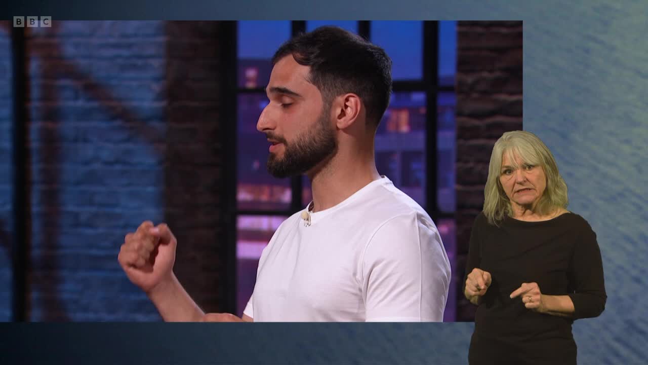 A young man in a white t-shirt gestures with his left hand, his brow furrowed in concentration, as a woman with grey hair signs to his right. This is part of a BBC Two broadcast.