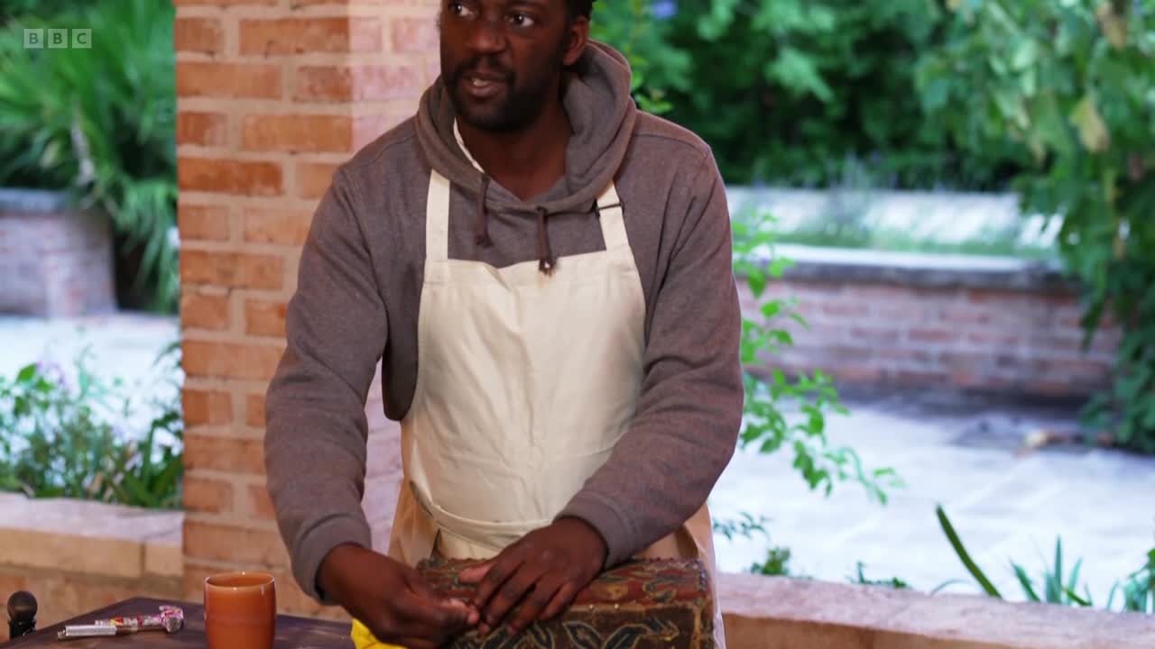 A man in an apron works on a decorative box. He's on a patio, with greenery behind him, likely for a BBC Two programme.