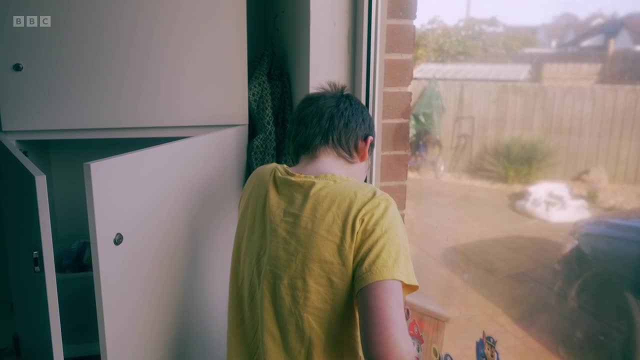 A young person in a yellow t-shirt stands by a window, looking out at a garden. A BBC Two logo is visible in the top left corner.