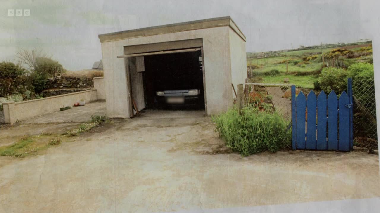 A silver car is parked inside an open garage. Beyond the garage, a bright blue gate stands beside a grassy hillside.