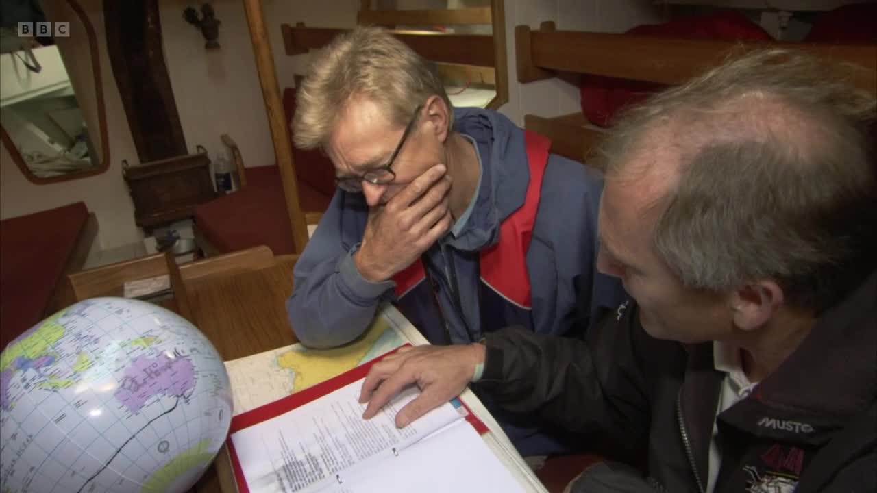 Two men pore over charts and a globe inside a boat's cabin. One man, wearing glasses, rests his chin on his hand as he studies the documents.